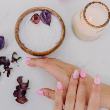 Close up of a pair of hands with pink manicured nails rubbing eachother, a pot of sugaring scrub next to them, with some scattered flowers, links to Sugaring Services at Athena's Glow Salon